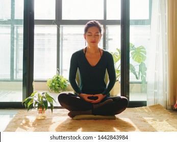 Pretty Chinese Young Woman Meditating At Home, Sitting On Floor With Furry Cushion In Sun Light, Exercise, Lotus Pose, Prayer Position, Namaste, Working Out, Feeling Peace And Wellness Concept.