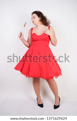 pretty chubby fashion girl wearing red pinup dress and posing with tasty colorful lollipop on white studio background