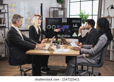 Pretty concentrated blond female boss leader at a meeting together with her diverse multiracial colleagues, in modern boardroom with big TV plasma screen, presenting world map and charts. - Powered by Shutterstock