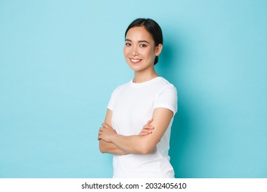 Pretty confident young asian woman looking upbeat, cross arms chest and smiling, standing half-turned over blue background with determined expression, concept of lifestyle and beauty