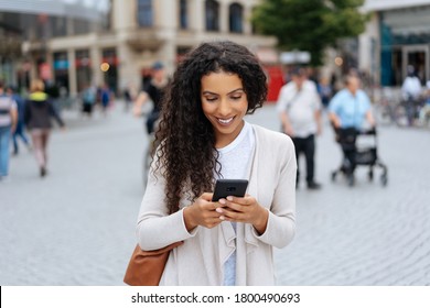 Pretty Curly Brunette Woman In Casual Clothes Smiling While Texting On Mobile Phone In Her Hands, Standing In The Middle Of A Crowded City Square. Front Portrait With Blurred Background