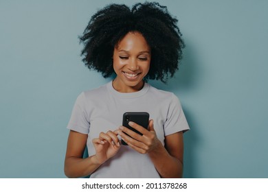 Pretty curly hair woman holds modern mobile phone, types messages on smartphone device, enjoys online communication, downloads special app for chatting, smiles tenderly, isolated on blue studio wall - Shutterstock ID 2011378268