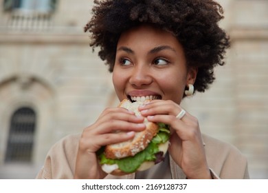 Pretty curly haired woman bites delicious sandwich poses outdoors at street looks away dressed casually has quick snack while walking outside being hungry. People lifestyle and fast food concept - Shutterstock ID 2169128737