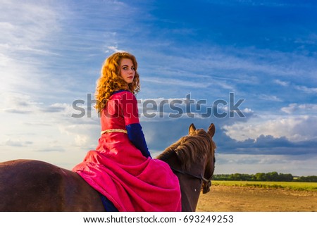A pretty curly red haired attractive young Caucasian white woman looks at camera top down, riding a horse turns holding reins in sky clouds fields horizon. Horizontal day, summer outdoors copy-space