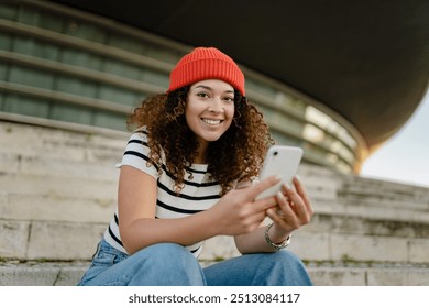 pretty curly smiling woman sitting in city street in striped t-shirt and knitted red hat, using smartphone, smiling happy - Powered by Shutterstock