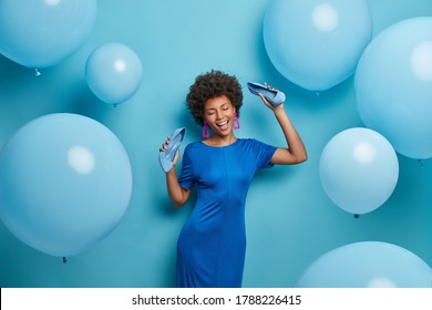 Pretty curly woman chills at party, dances happily, keeps hands raised with shoes, spends time in night club, takes off high heel shoes, poses against blue wall. Monochrome shot. Holiday, celebration