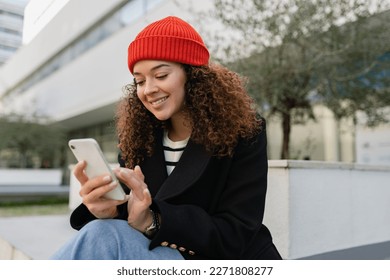 mujer bonita y rizada caminando en la calle de la ciudad con un elegante abrigo de otoño, usando un teléfono inteligente, usando sombrero de punto rojo