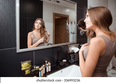 Pretty curly young female looking in mirror while brushing hair standing in bathroom 