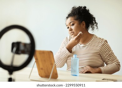 Pretty curly young hispanic woman in casual outfit sitting at vanity table in front of mirror, using face toner cleansing face with micellar water, recording video for followers at home, copy space - Powered by Shutterstock