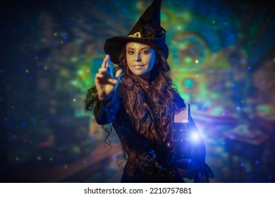 A Pretty Curly-haired Witch In A Black Hat And Black Dress With A Magic Book Of Spells Reaches Hand Towards The Camera And Smiles. Wonderful World Of Magic. Halloween. 