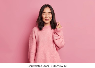 Pretty Dark Haired Woman With Gentle Smile Makes Korean Like Sign Expresses Love To You Wears Casual Long Sleeved Jumper Isolated Over Pink Background. Mini Heart Gesture. Body Language Concept