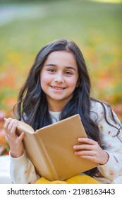 A Pretty Dark-haired 10-year-old Girl In A Knitted Sweater Reads A Book Sitting In A Clearing In An Autumn Park. Picnic In Nature. Happy Child Is Learning. Fall.