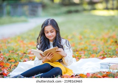 A Pretty Dark-haired 10-year-old Girl In A Knitted Sweater Reads A Book Sitting In A Clearing In An Autumn Park. Picnic In Nature. Happy Child Is Learning. Fall.