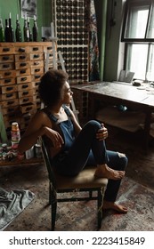 Pretty And Dreamy African American Artist In Overalls Sitting Barefoot In Workshop And Looking Away Through Window