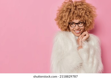 Pretty dreamy fashionable woman with curly hair wears spectacles ad fur white coat keeps hands near face looks gently away isolated over pink background blank copy space for your advertisement