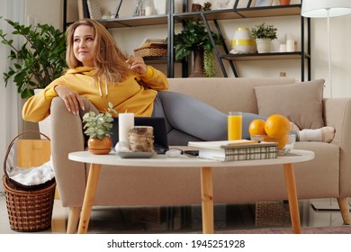 Pretty Dreamy Young Plus Size Woman Relaxing On Sofa At Home, Watching Show On Digital Tablet And Drinking Orange Juice