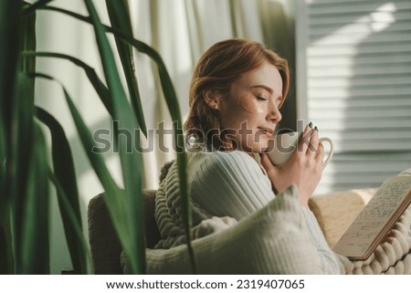 Pretty dreamy young woman sitting on couch covered with a blanket, drinking coffee indoors at home room. Cosy scene, cozy home concept