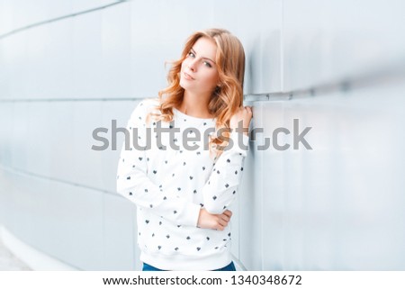 Pretty elegant positive young blond woman with a beautiful smile in a fashionable pullover near a white modern wall indoors. Stylish positive girl model on vacation. Spring style youth clothing.