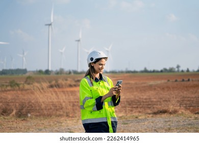 Pretty engineer or technician woman with safety protection equipment stand with using mobile phone and stand in front of windmill cluster in the field. - Shutterstock ID 2262414965
