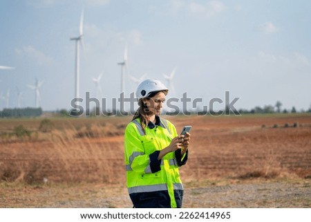 Pretty engineer or technician woman with safety protection equipment stand with using mobile phone and stand in front of windmill or wind turbine cluster in the field.