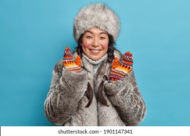 Pretty Eskimo Woman Dresses For Cold Weather Smiles Happily Wears Fur Coat And Hat Raises Hands In Mittens Has Good Mood Isolated Over Blue Background. Asian Female From Tundra In Winter Clothes.