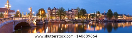 Pretty evening illuminations of the historic and richly decorated Blauwbrug (1883) bridge in Amsterdam. Skinny bridge and Stopera music theatre visible. Stitched panoramic image.