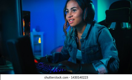 Pretty and Excited Black Gamer Girl in Headphones is Playing First-Person Shooter Online Video Game on Her Computer. Room and PC have Colorful Neon Led Lights. Cozy Evening at Home.