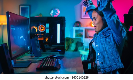 Pretty And Excited Black Gamer Girl In Headphones Is Winning In First-Person Shooter Online Video Game On Her Computer And Puts Hands Up. Room And PC Have Colorful Neon Led Lights Cozy Evening At Home