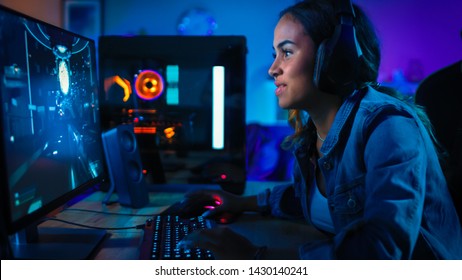 Pretty And Excited Black Gamer Girl In Headphones Is Playing First-Person Shooter Online Video Game On Her Computer. Room And PC Have Colorful Neon Led Lights. Cozy Evening At Home.