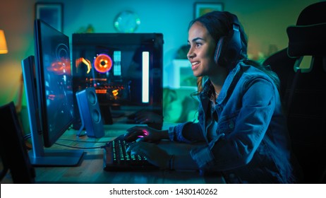 Pretty and Excited Black Gamer Girl in Headphones is Playing First-Person Shooter Online Video Game on Her Computer. Room and PC have Green Neon Led Lights. Cozy Evening at Home.