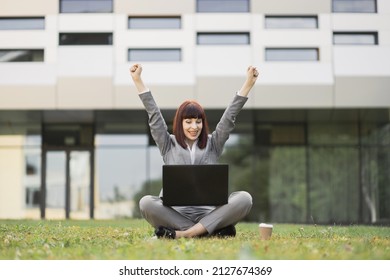 Pretty excited woman in business suit with raised hands, sitting crosslegged on green grass in front of office, using a laptop, celebrating success, screaming and raising arms. Well done, good news