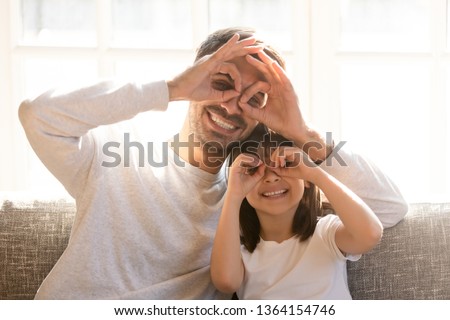 Pretty family father adorable daughter sitting on sofa do funny faces making with fingers eyewear shape like glasses looking through binoculars, have fun with child free time play tricks fool concept