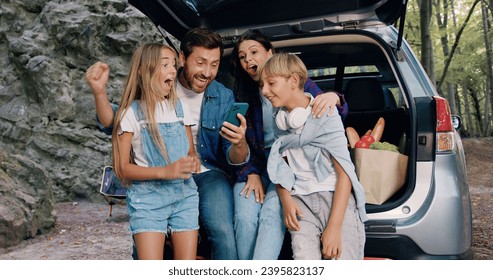 Pretty family mother father and children sitting in car with trunk open laughing browsing online swiping on smartphone. Happy family with their son and daughter using phone sitting in trunk of car - Powered by Shutterstock