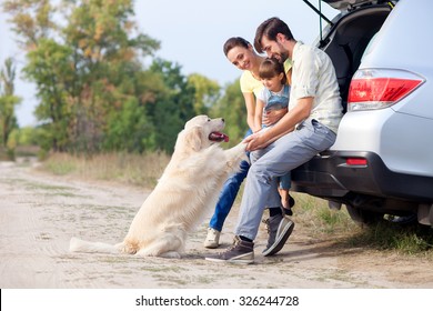 Pretty Family Is Resting In Forest And Playing With Dog. They Are Sitting On Open Car Boot And Embracing. The Man And Woman With Girl Are Smiling. Copy Space In Left Side