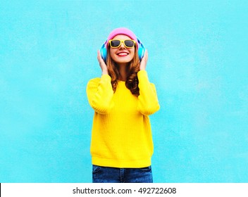 Pretty Fashion Cool Smiling Girl Listening To Music In Headphones Wearing A Colorful Pink Hat, Yellow Sunglasses And Sweater Over Blue Background