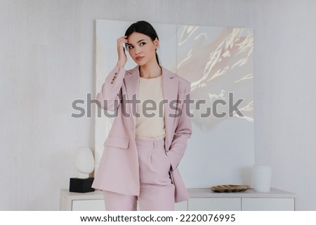 Pretty fashion model posing touching hair dressed in light pink suit standing at living room. Attractive asian young adult woman looking aside. Gorgeous elegant Korean girl preparing to interview.