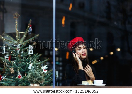 Pretty fashion young woman sitting in the cosy cafe and talk on the phone. Christmas tree on the background. Red hat nad lips. Winter holiday concept