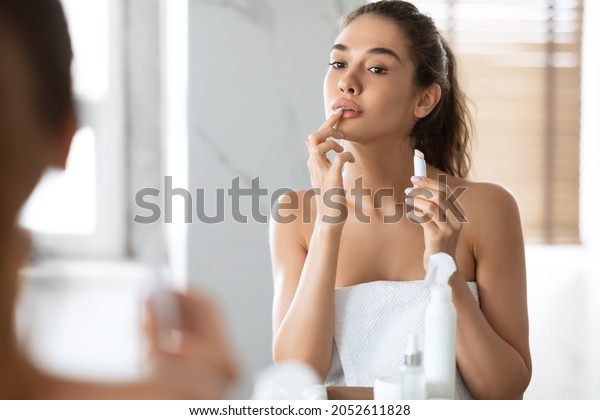Pretty Female Applying\
Lip Balm Moisturizing Caring For Skin Standing Near Mirror In\
Modern Bathroom At Home. Makeup And Cosmetics, Lips Skincare\
Concept. Selective Focus