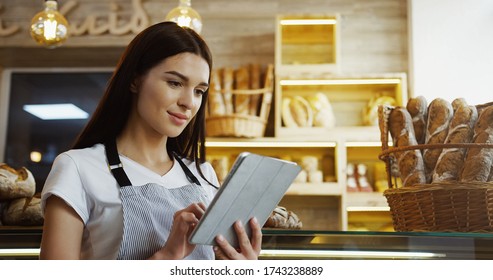 Pretty female baker working in bakery shop using digital tablet computer while standing at counter.