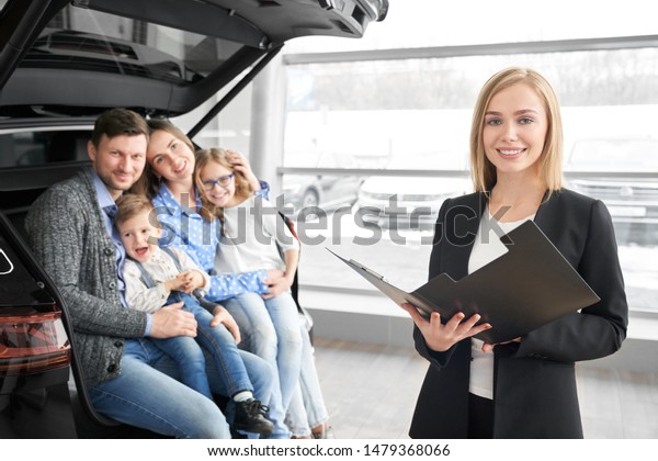 Pretty female car dealer looking at camera, posing\
and keeping contract in auto selling to lovely family. Happy client\
sitting in car at background and enjoying purchasing. Concept of\
auto salon.