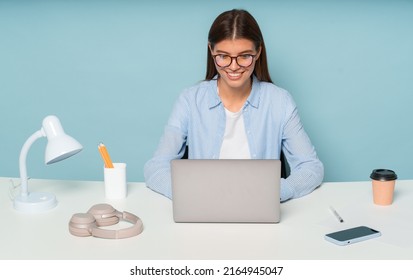 Pretty female copywriter in glasses working on new article using laptop and wireless connection sitting at table surrounded with office supplies and phone with copy space screen, isolated on blue
