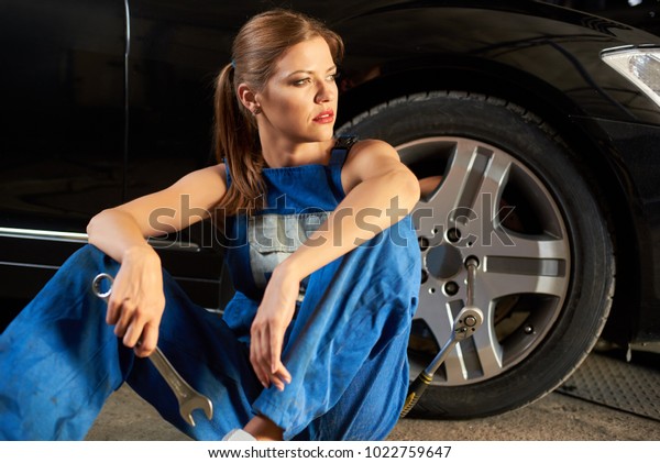Pretty female\
mechanic sits near the wheel of black car. Girl is dressed in work\
uniform and holds a wrench in her hands. In the wheel you can see\
the key for removing the\
wheel