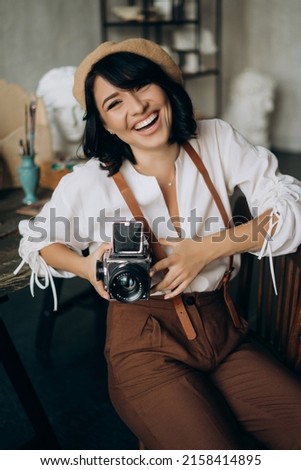 Pretty female sculptor 
with beret and suspenders holding retro camera (authoring photo)