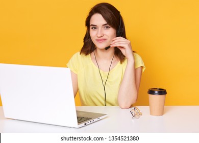 Pretty female sits at white desk with opened laptop computer, writes email, uses high speed internet, poses isolated over yellow background, works or studies online. People and technology concept.