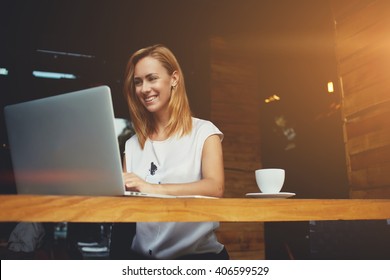 Pretty Female Student With Cute Smile Keyboarding Something On Net-book While Relaxing After Lectures In University, Beautiful Happy Woman Working On Laptop Computer During Coffee Break In Cafe Bar