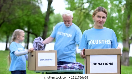 Pretty female volunteer holding box, activists sorting clothes on background - Powered by Shutterstock