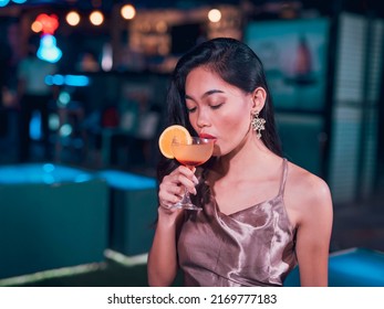 A pretty Filipino woman in a shiny dress drinks a Tequila Sunrise cocktail. A single and attractive woman enjoying an alcoholic drink at a bar. Nightlife scene. - Powered by Shutterstock