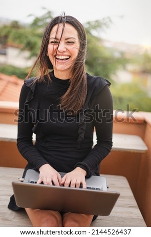 Pretty forty woman smile keeping a laptop computer on her knees and typing on the keyboard sitting on the balcony