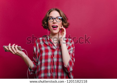 Pretty funny girl with dark hair listening music in white earphones and looking away. Indoor photo of charming young woman in trendy glasses enjoying song on claret background.