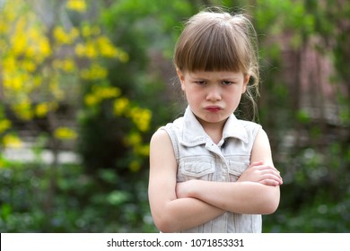 Pretty funny moody little blond preschool girl in white sleeveless dress looks into camera feeling angry and unsatisfied on blurred summer background. Children tantrum concept.